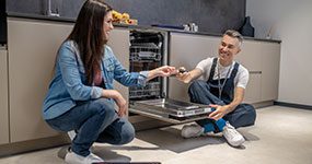 A man and woman sitting on the floor near an oven.